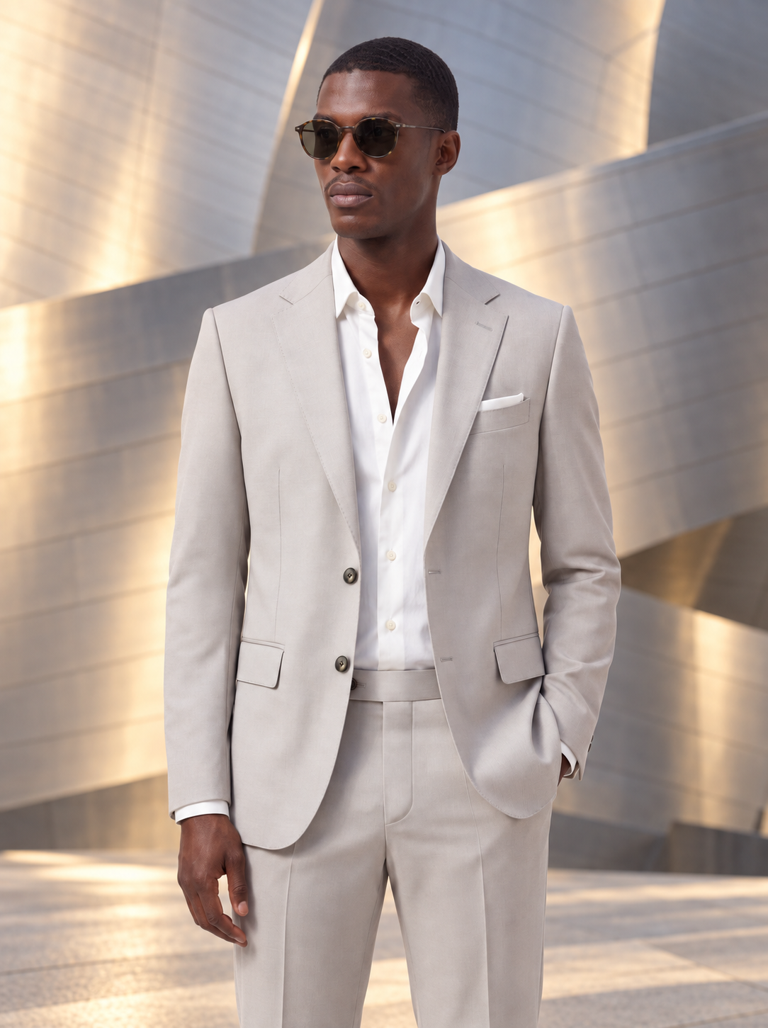 A man wearing a light beige two-piece suit with a white open-collar dress shirt and sunglasses, standing in front of a modern metallic building.