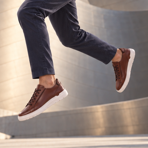 A man wearing a white long-sleeve shirt, dark gray trousers, and brown slip-on sneakers jumps in front of the modern metallic architecture of the Walt Disney Concert Hall in Los Angeles.