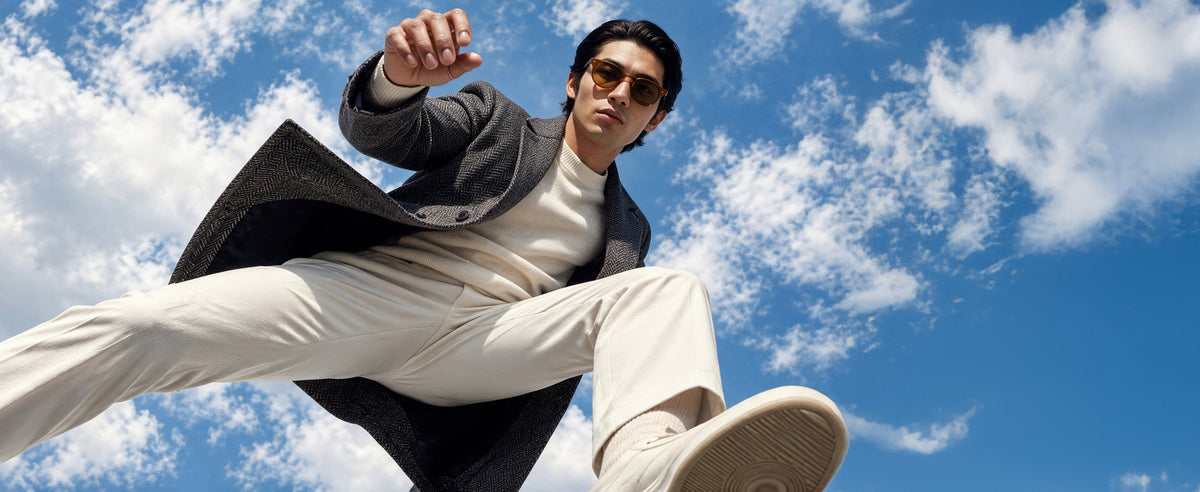 A man in a dark blazer, white shirt, and white trousers jumps against a bright blue sky with white clouds.
